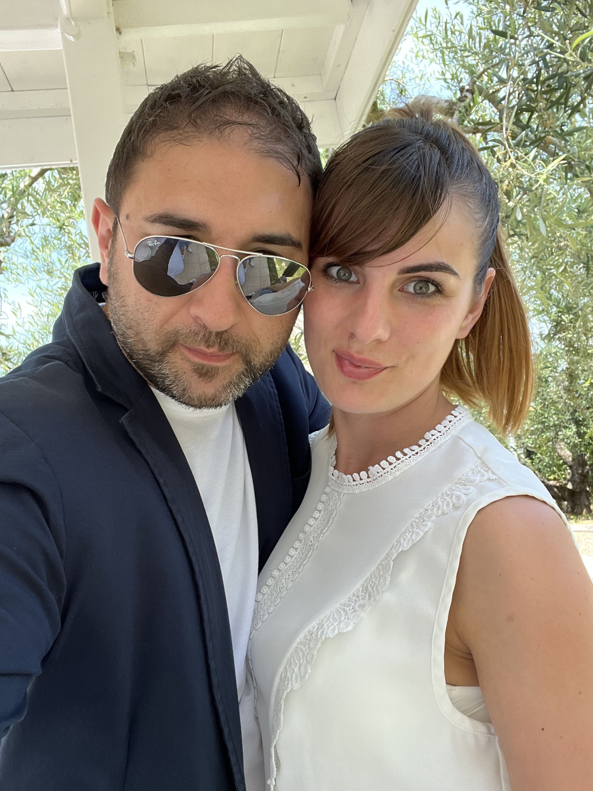 Due persone in posa ravvicinata all'aperto, una con occhiali da sole e blazer scuro, l'altra con un abito bianco.