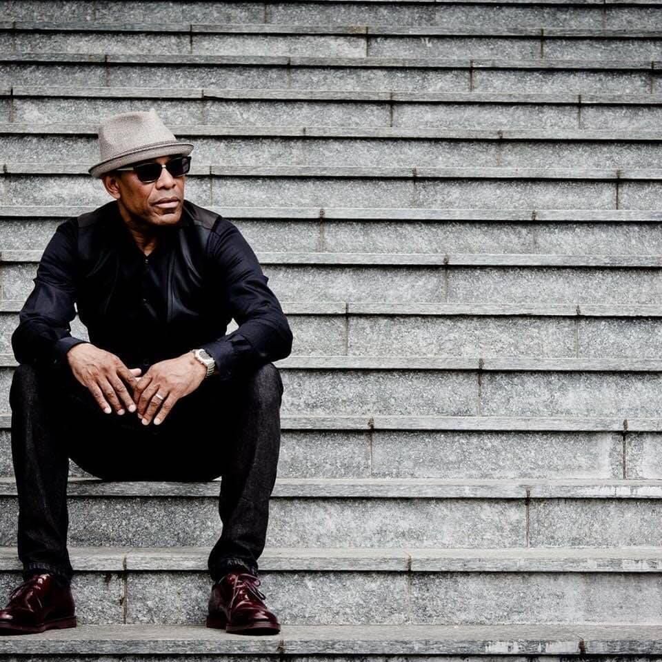 Un uomo vestito di nero, con cappello e occhiali da sole, è seduto su dei gradini di pietra e indossa scarpe rosse.