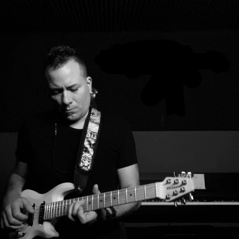 Persona che suona la chitarra elettrica in uno studio buio, con indosso una camicia nera e una tracolla fantasia.