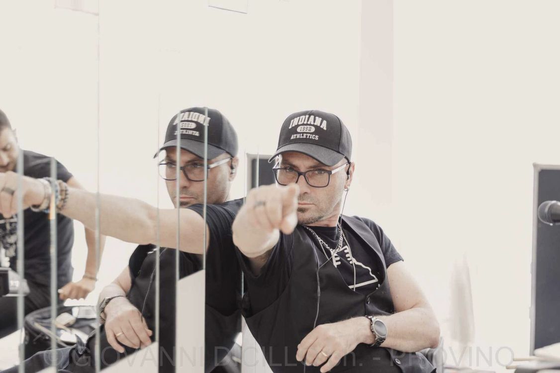Tre uomini con berretti neri siedono dietro le sbarre, uno dei quali indica verso la telecamera.