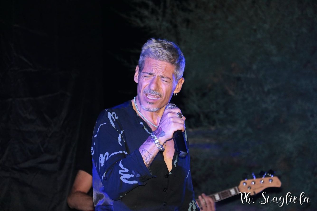 Cantante che si esibisce sul palco con un microfono, indossando una camicia scura sotto luci blu-violacee.