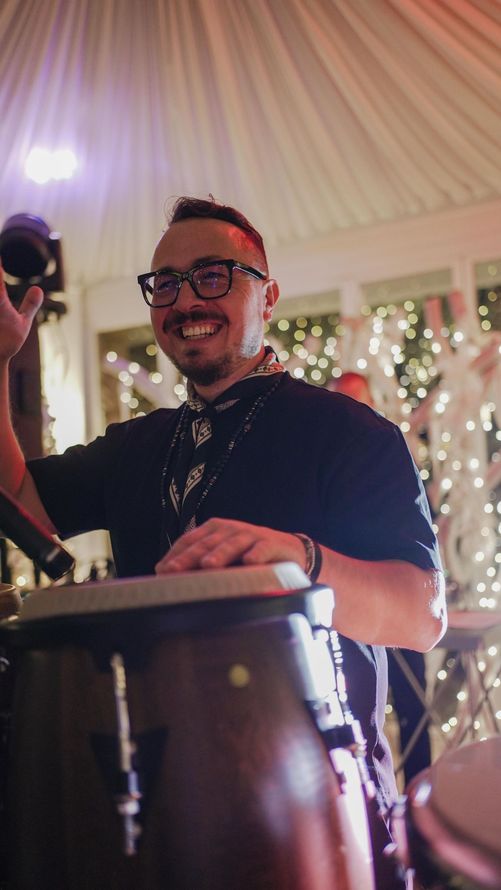 Un batterista sorridente dietro un tamburo conga durante un evento illuminato da luci festive.
