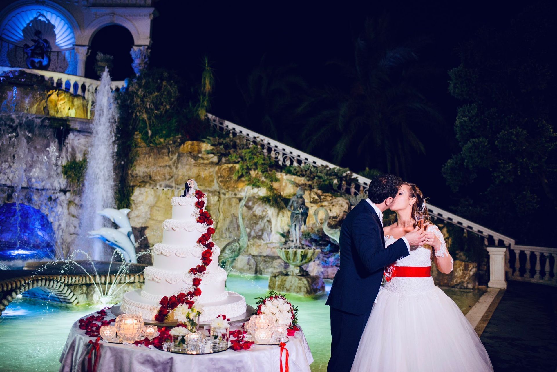 Gli sposi si scambiano un bacio accanto a una torta nuziale illuminata e a una fontana durante il ricevimento serale.