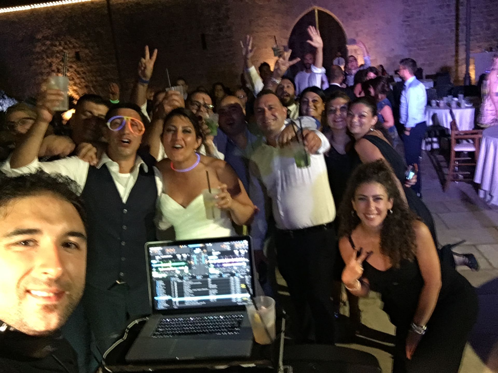 Gruppo di persone in posa a una vivace festa al chiuso, sorridenti e con in mano dei drink, attorno a una postazione da DJ con un laptop.