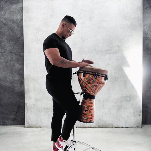 Un uomo in piedi suona un djembe decorato in uno studio dall'atmosfera minimalista.