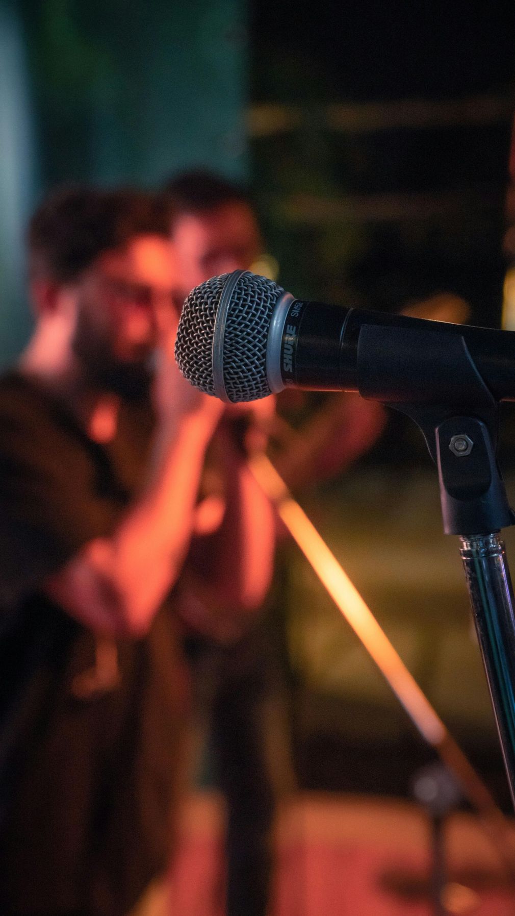 In primo piano, un microfono su un supporto, con sullo sfondo una cantante sfocata e le luci del palcoscenico.