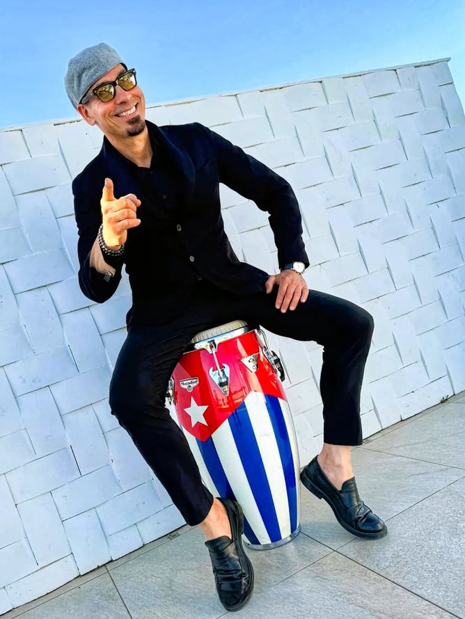 Un uomo vestito di nero è seduto su un tamburo conga rosso, bianco e blu, appoggiato a una parete bianca, mentre sorride e gesticola.