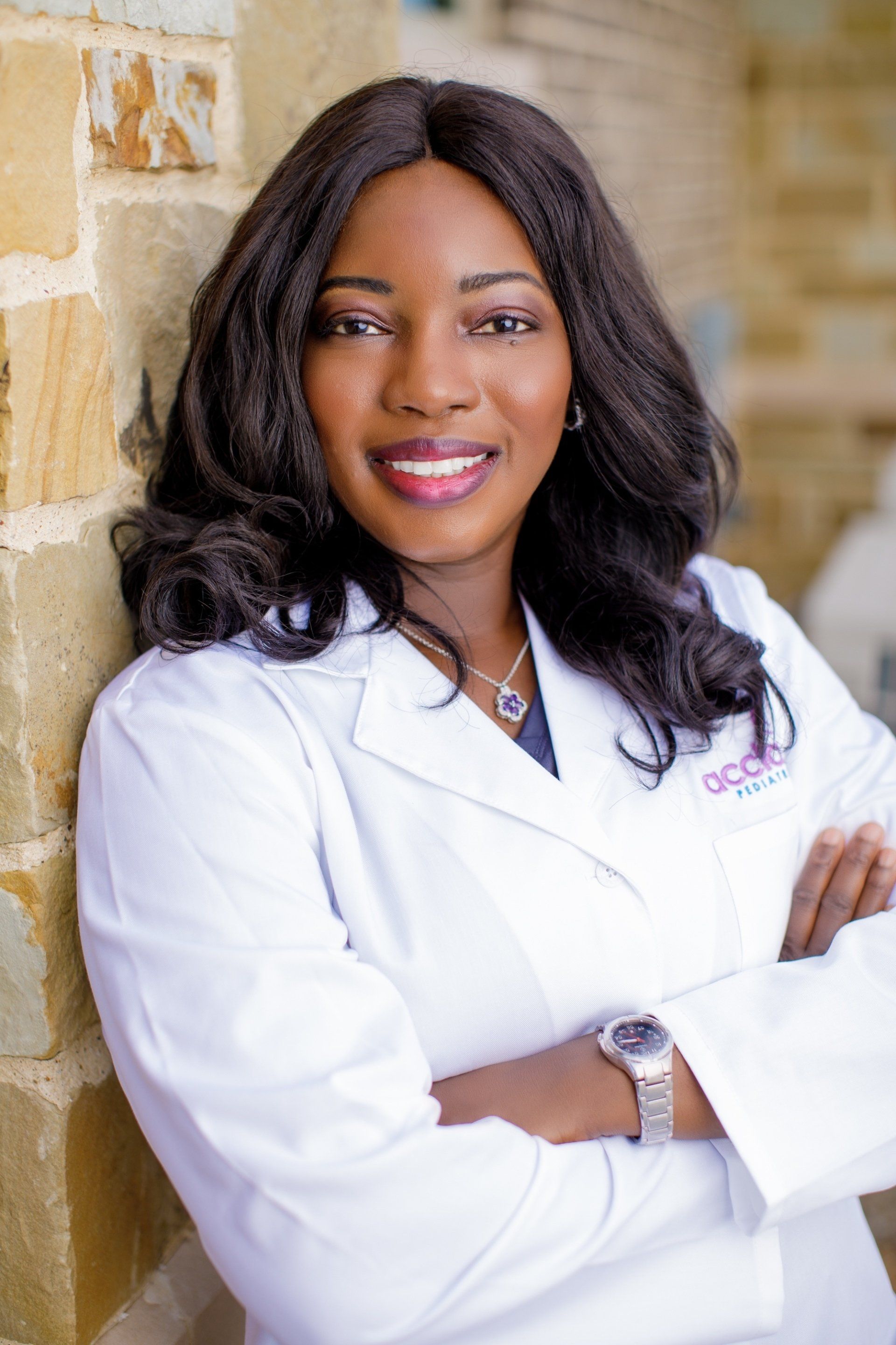 A woman in a white lab coat is standing in front of a brick wall with her arms crossed.