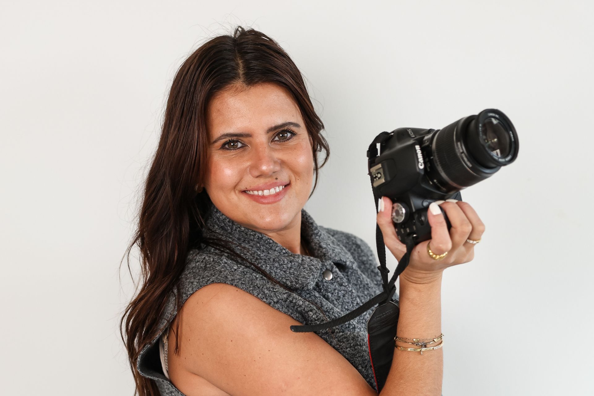 Vrouw met een spiegelreflexcamera in haar handen, lachend naar de kijker; ze draagt een grijs vest en staat voor een witte achtergrond.