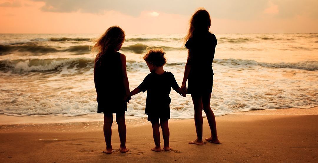 Three children are standing on a beach holding hands.