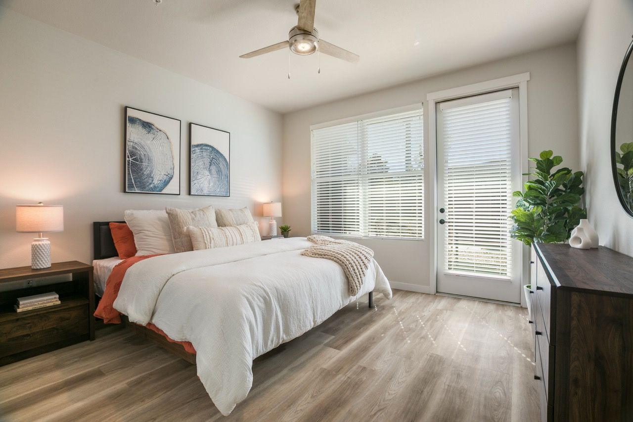 Bright modern bedroom in an apartment with a large bed, two nightstands, dresser, ceiling fan, and exterior door.