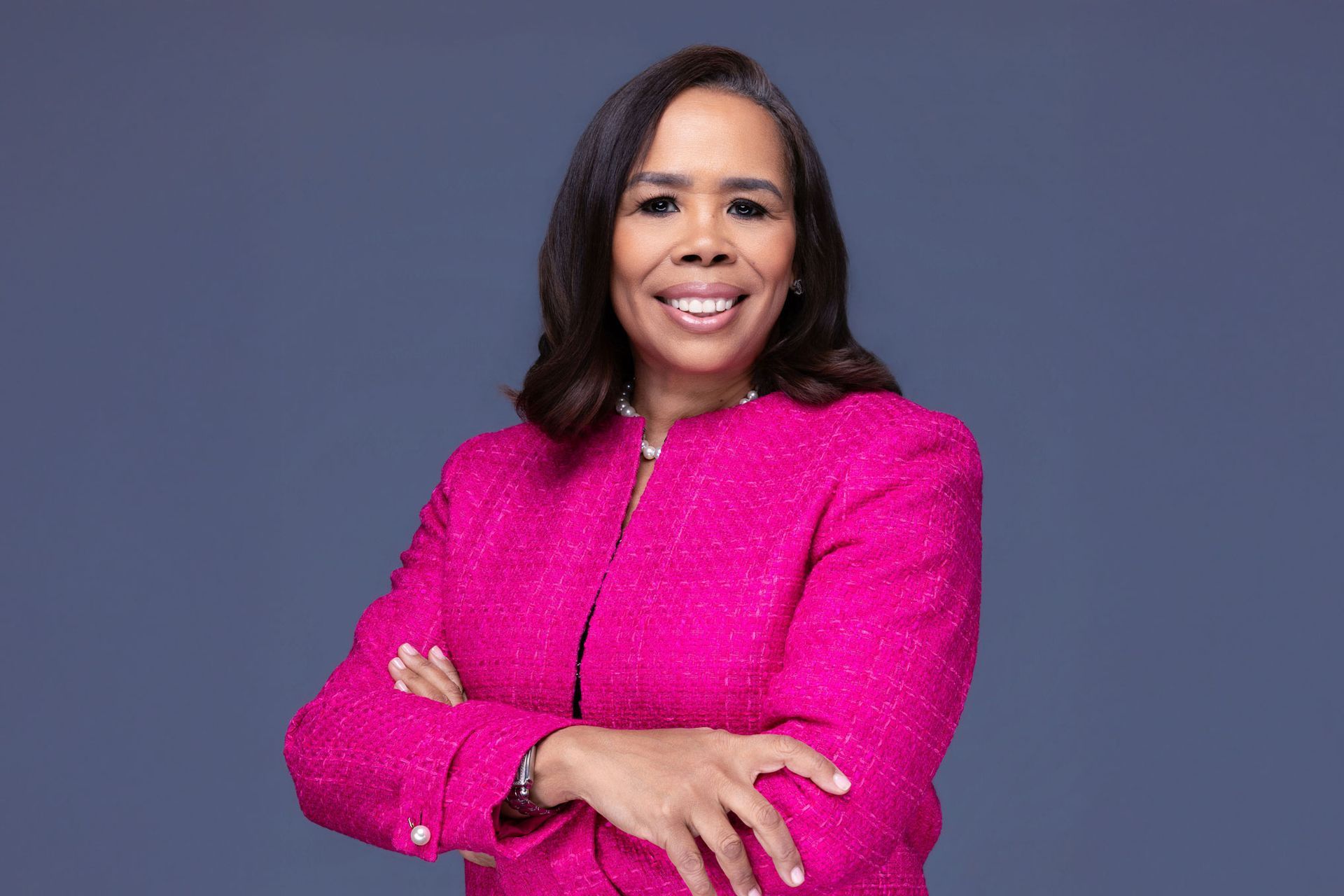 Andrea with arms crossed, wearing a bright pink textured jacket against a muted blue background.