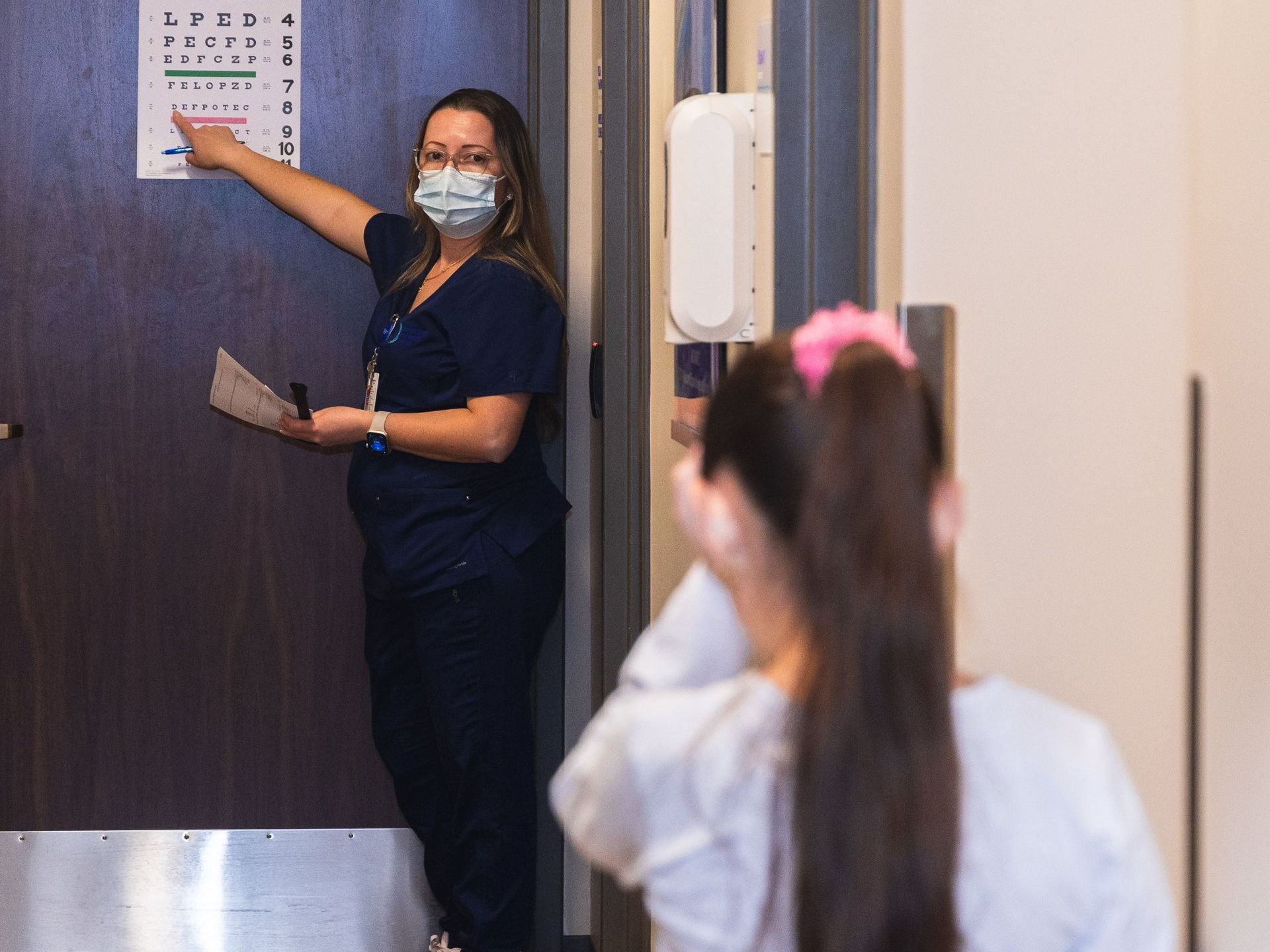 An image of a female doctor pointing to an eye chart in front of a little girl