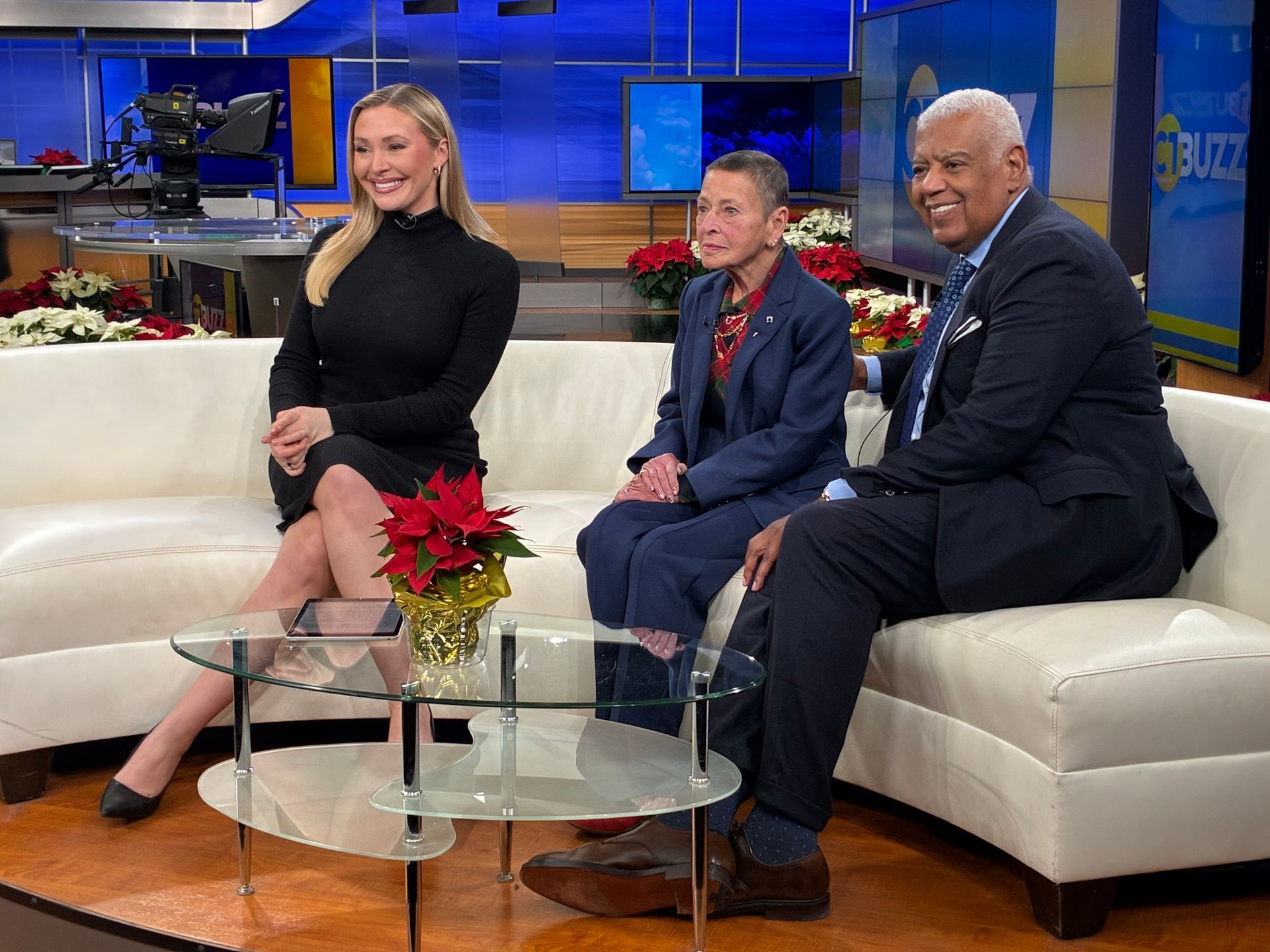Three people on a white couch in a TV studio. Two are wearing dark business attire; one is in a black dress.