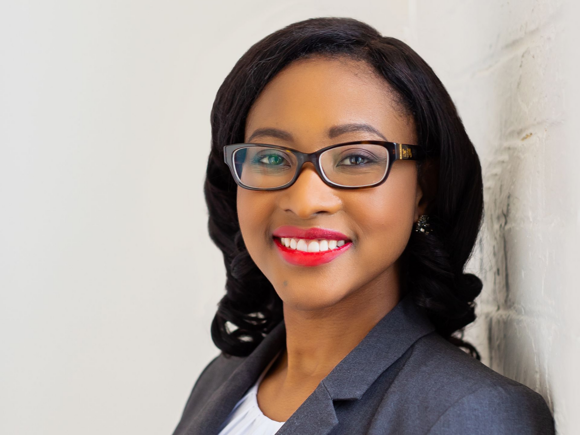 Woman with glasses and red lipstick smiles, wearing a gray blazer, leaning against a white wall.