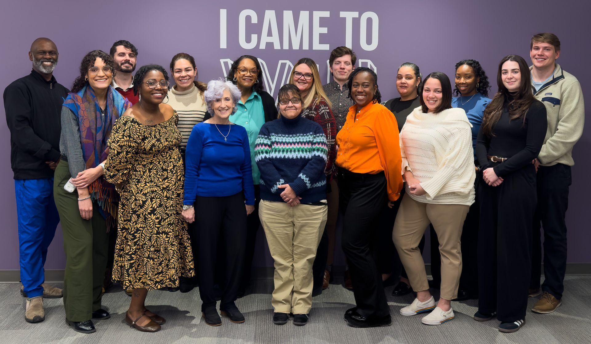 Group of people posing indoors in front of a purple wall with the words “I CAME TO” behind them.