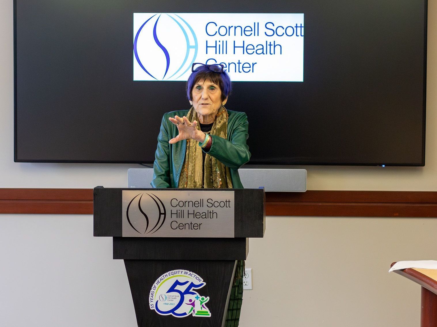 A woman is standing at a podium in front of a cornell scott hill health center sign.
