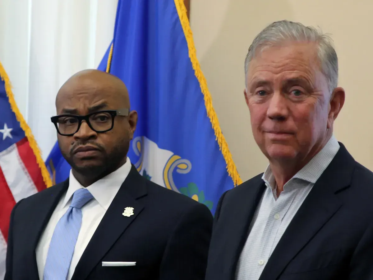 Two men in suits standing before flags, one wearing glasses and a lapel pin.