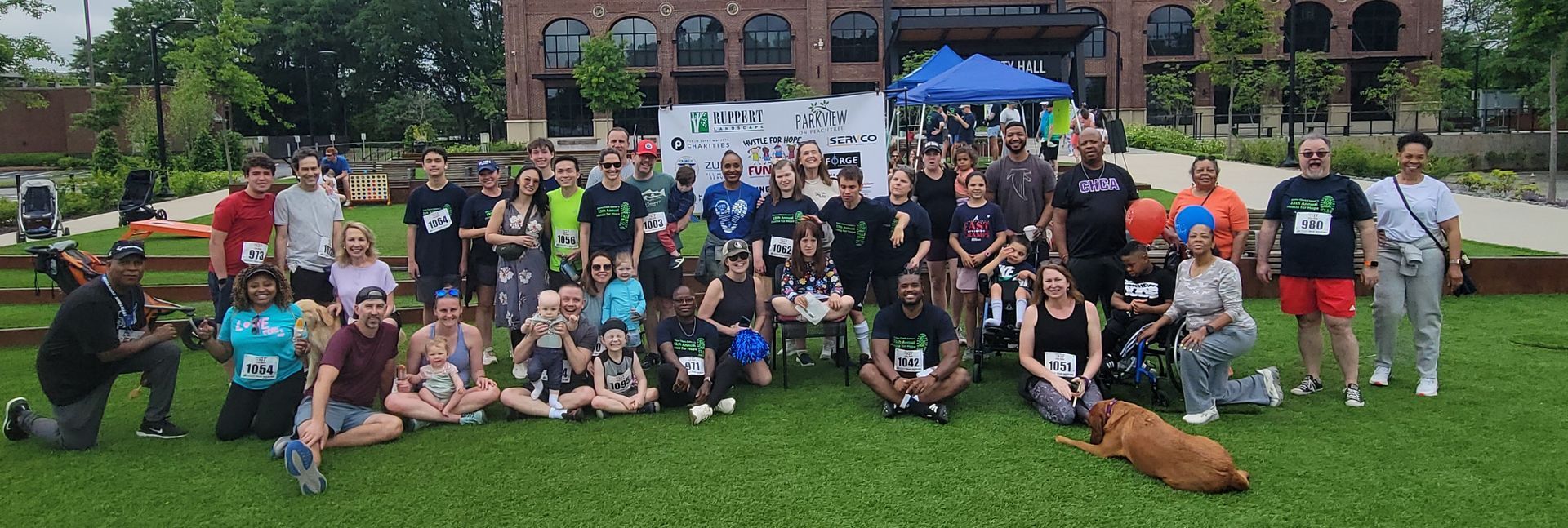 Group of people pose on grass with building backdrop, some wearing race bibs, event.
