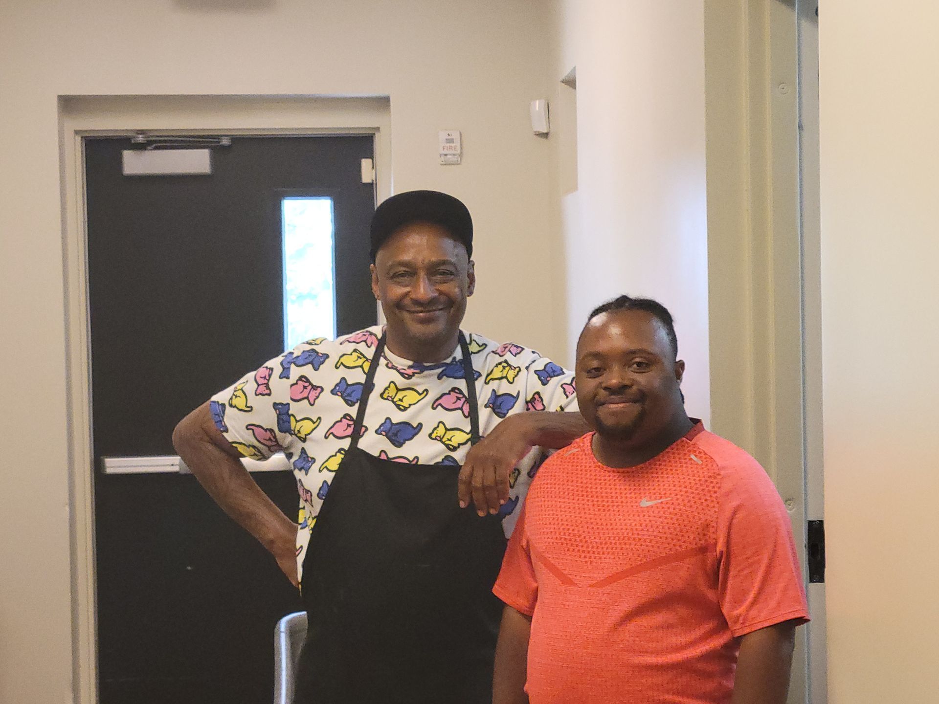 Two men smiling by a door. One in a colorful shirt and apron, the other in a red shirt. They are inside a building.