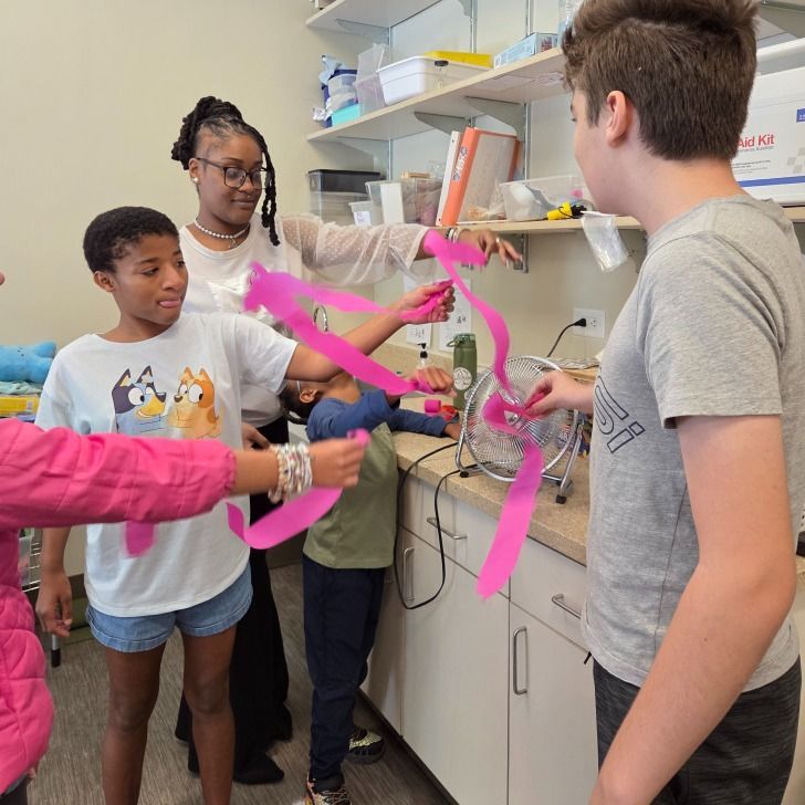Group of children and a teacher experiment with pink streamers near a fan.