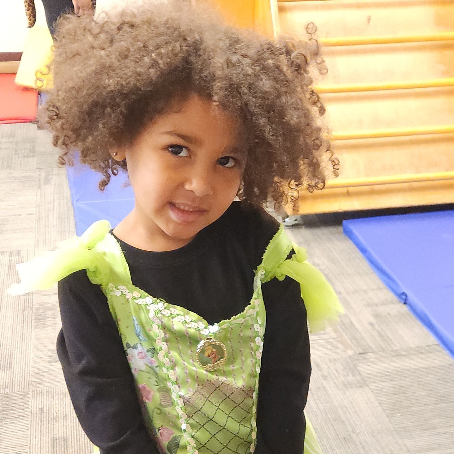Girl with curly brown hair, wearing a green and black costume, smiles indoors.