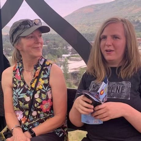 Two people in an enclosed cable car; woman with floral top and cap smiles, the other with black tee holds a brochure and speaks.
