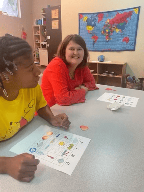 Two people sit at a table looking at papers. One smiles. A world map hangs in the background.