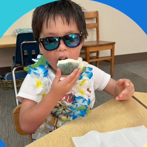 Boy wearing sunglasses and a lei, eating a cupcake, indoors.
