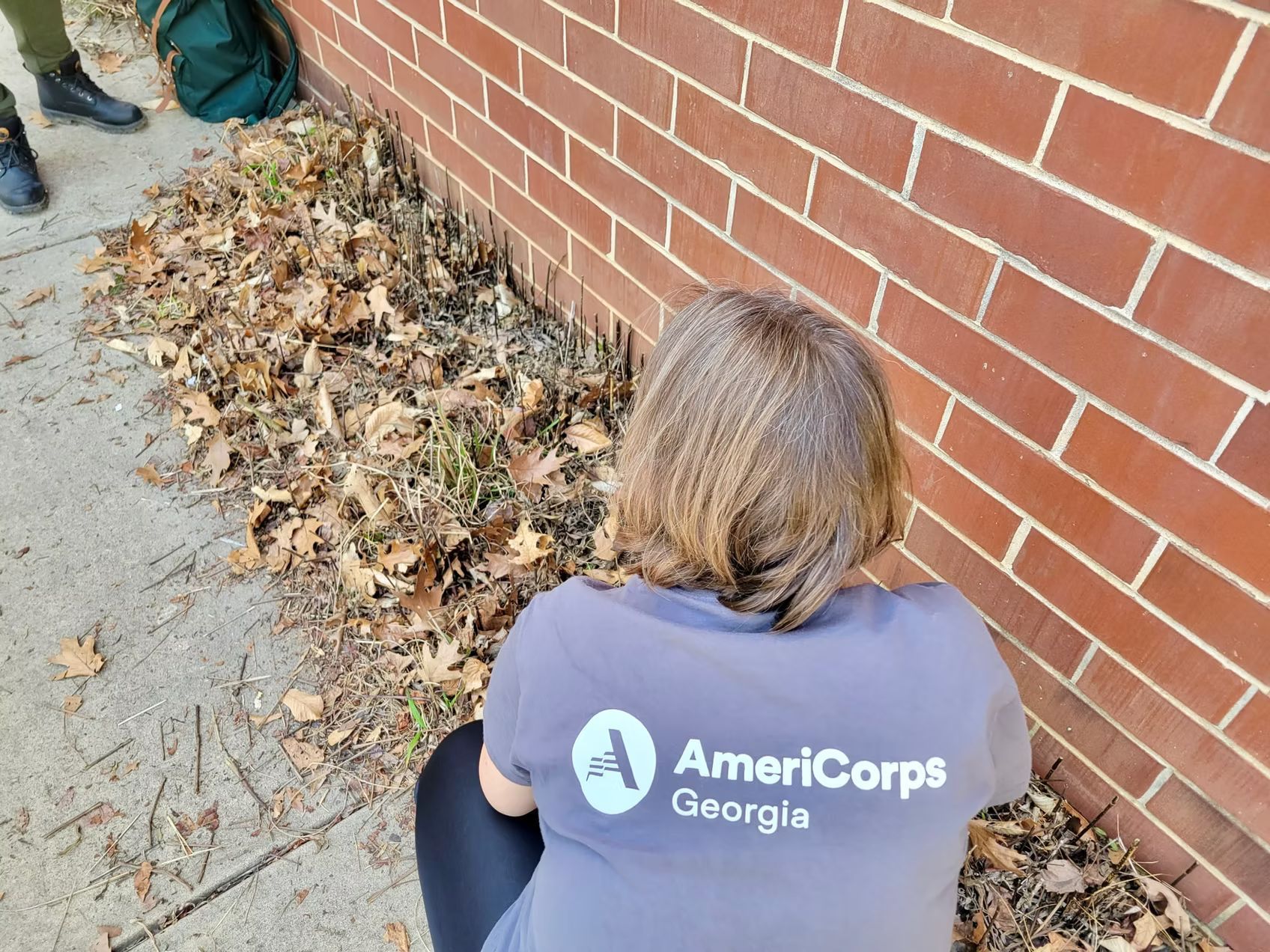 Person kneeling, wearing AmeriCorps shirt, near a brick wall and fallen leaves on the ground.
