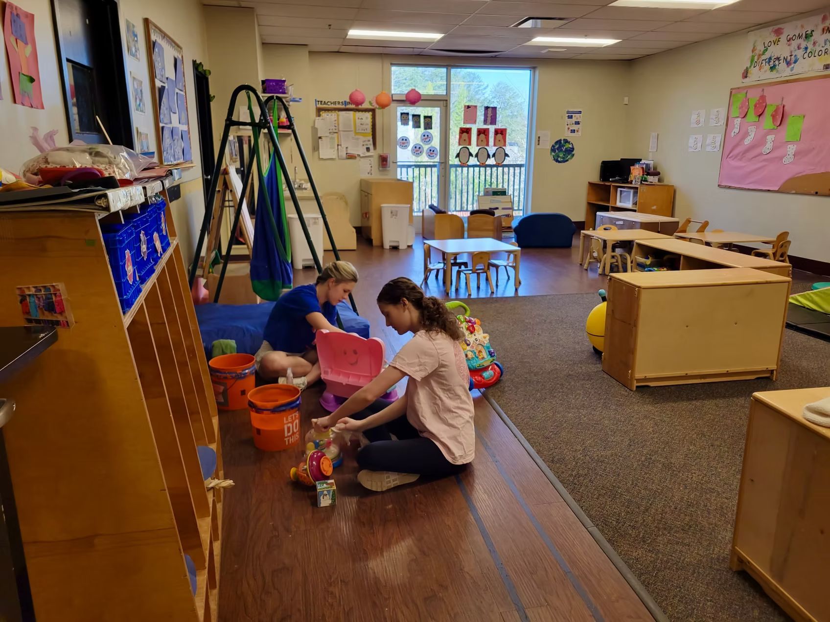Two people playing with toys on a playroom floor. Shelves and tables are visible.