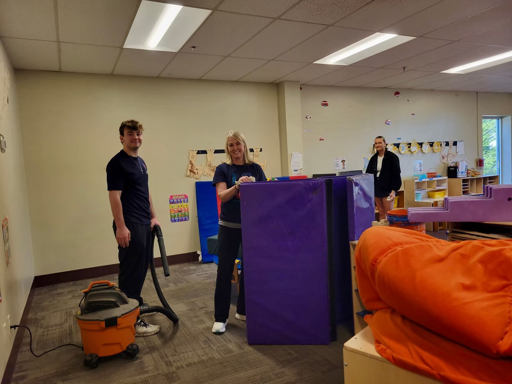 Three people clean a brightly lit room with a vacuum. One person holds the vacuum, another leans over large purple pads, the third stands in the background.