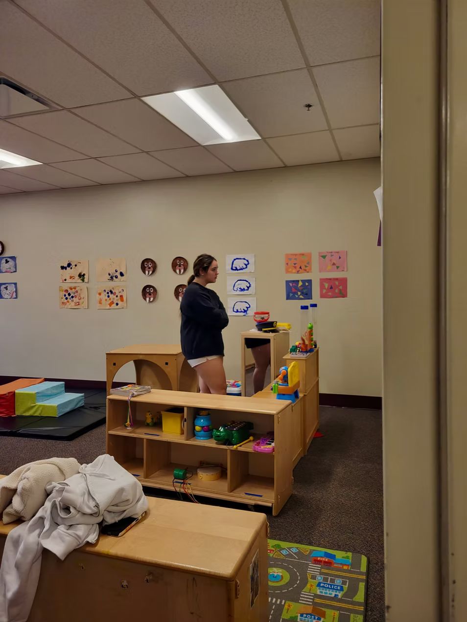 Woman in a children's play area with wooden shelves, artwork on the wall, and toys.
