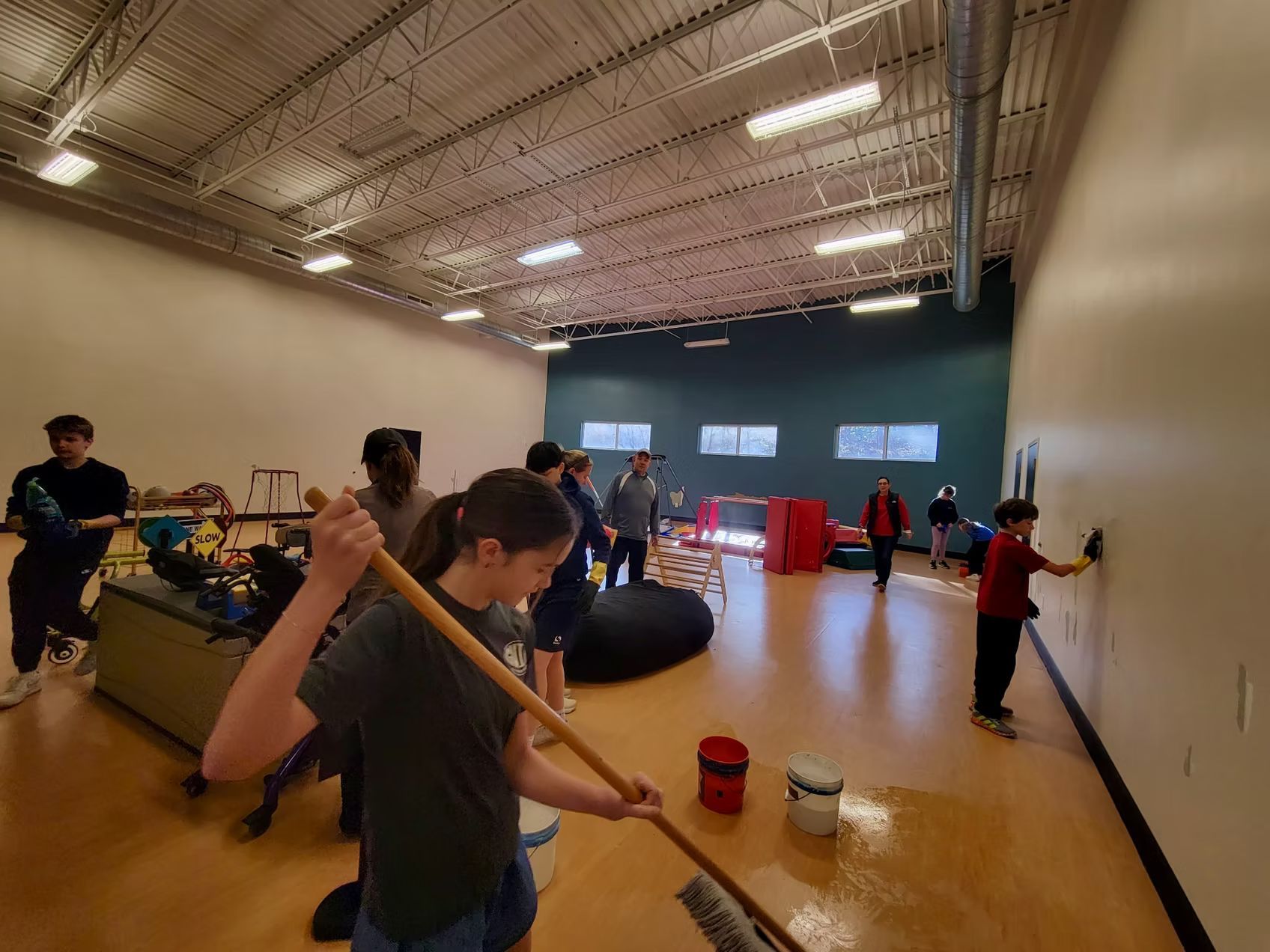 People painting an interior wall in a large room with wood flooring.