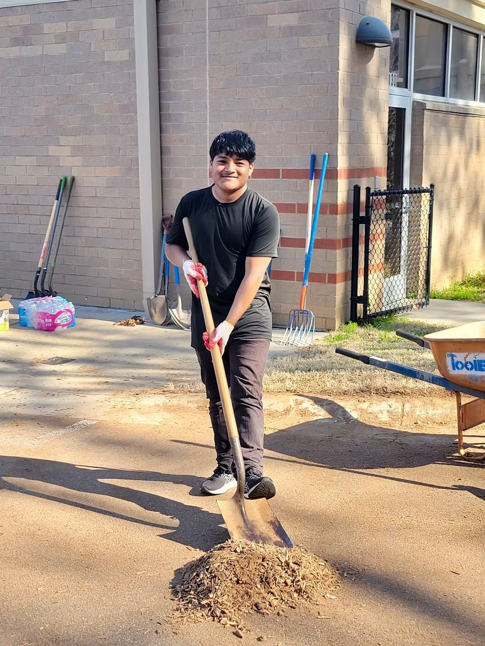 Person raking debris outside a building. He smiles, wearing a dark shirt and pants, with tools nearby.