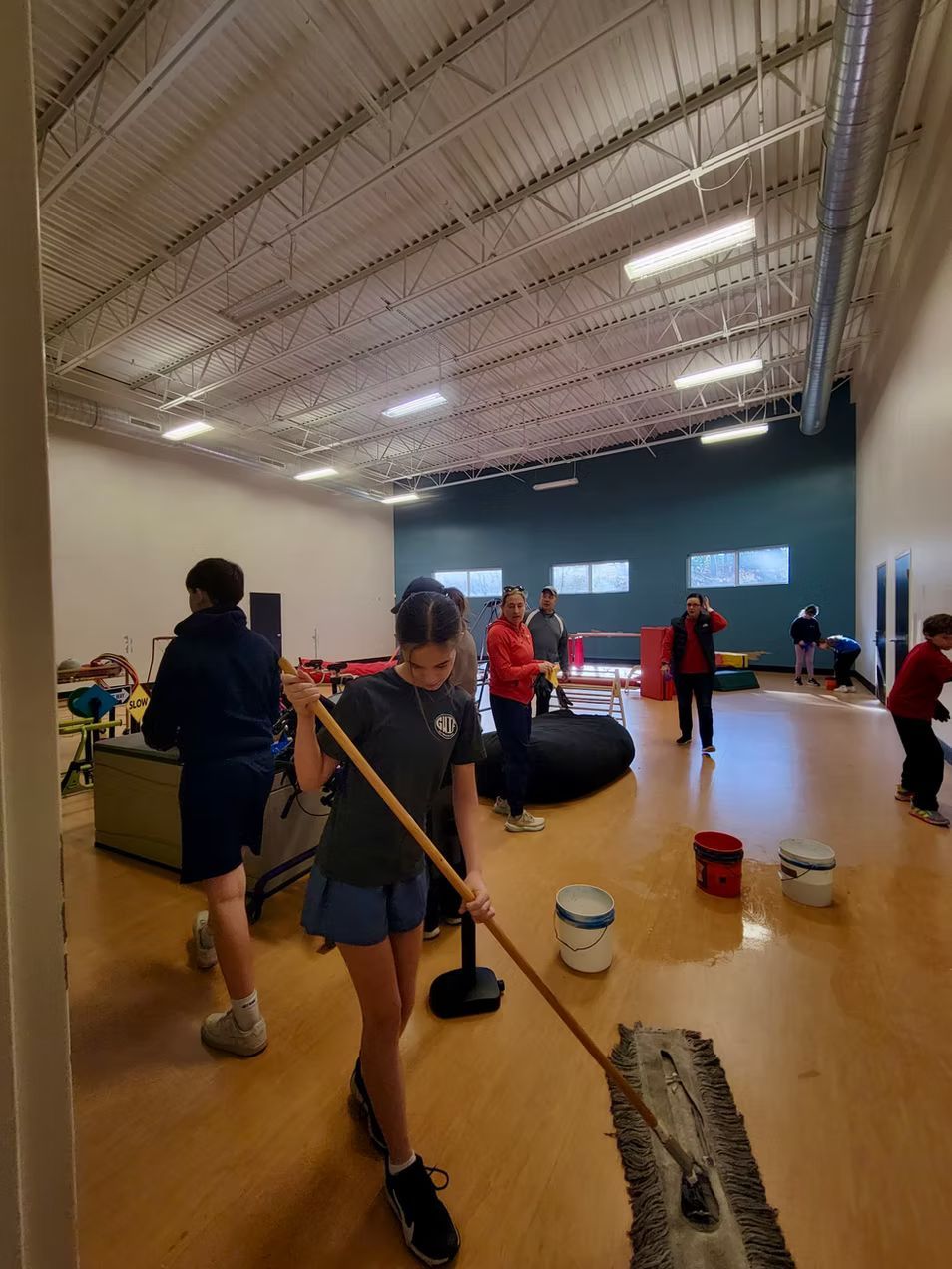 Person mopping a large room, other people in background. Light-colored floor, white ceiling, blue-green accent wall.