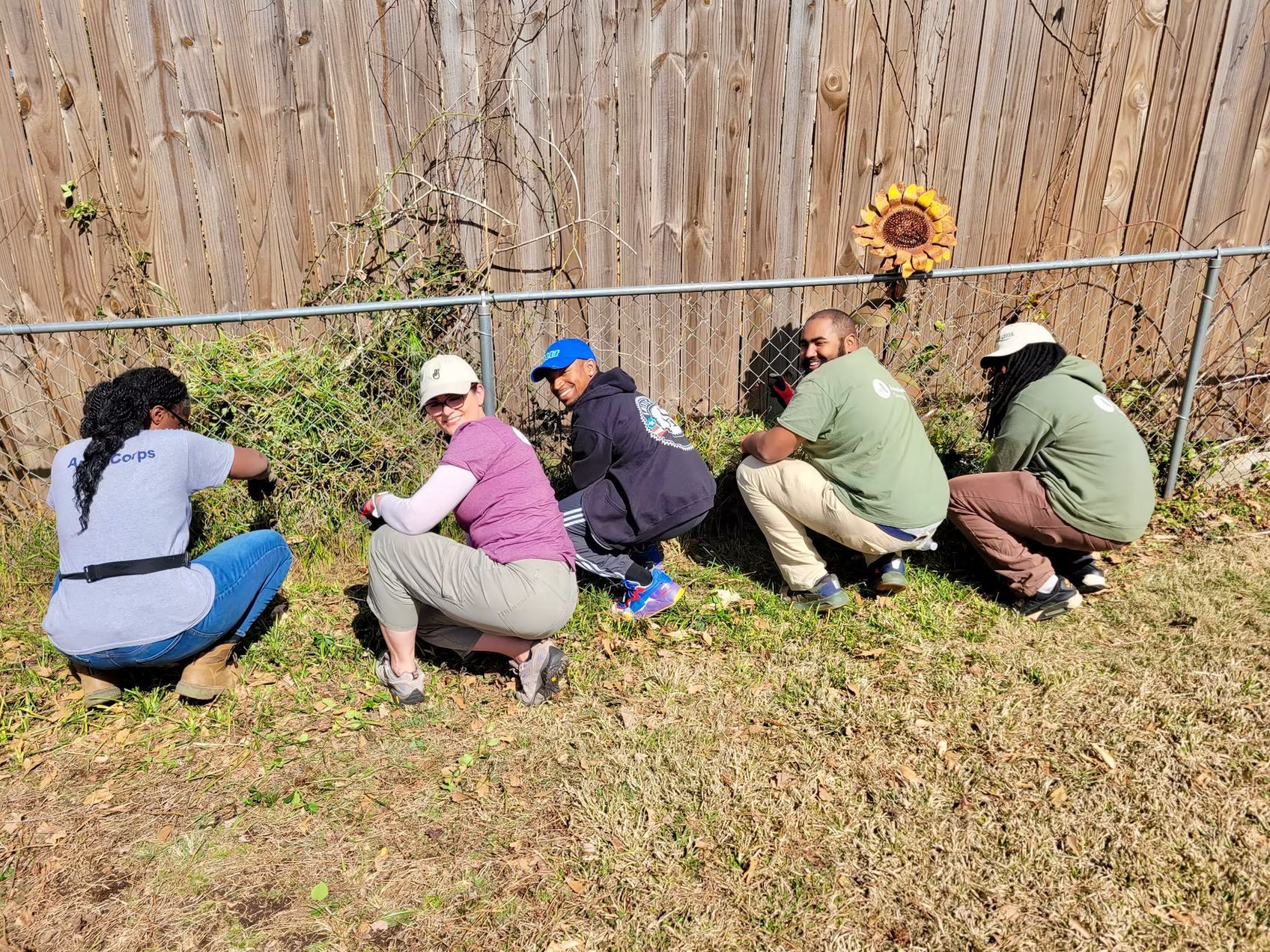 Five people crouched by a fence weeding a yard; sunflower decoration on fence.