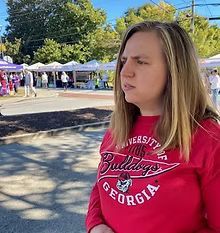 Woman in a red University of Georgia Bulldogs sweatshirt looks off-camera at an outdoor event with vendor tents in the background.