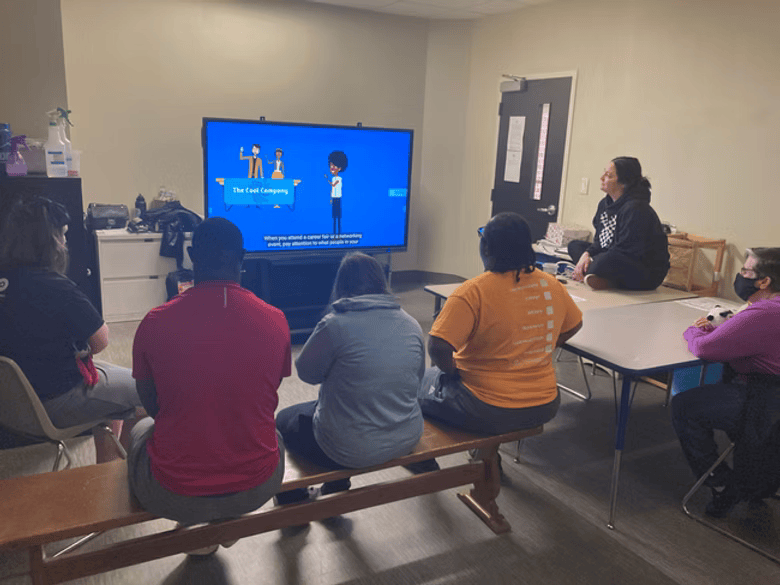 People watching a presentation on a large screen in a room. A person sits on a table, speaking.
