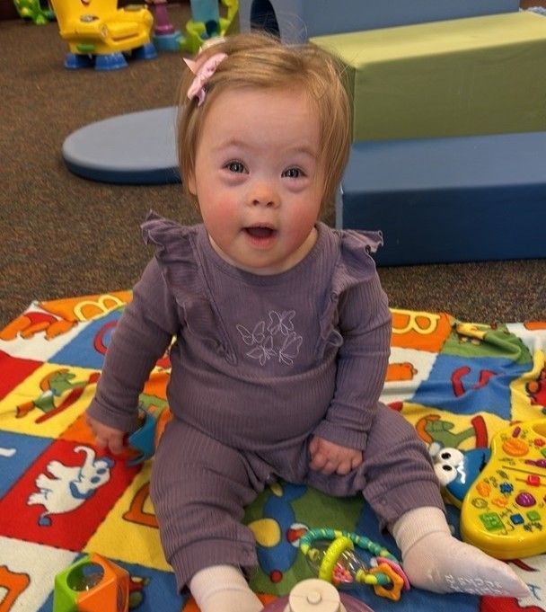 Child with Down syndrome in purple outfit sitting on a play mat with toys, smiling.