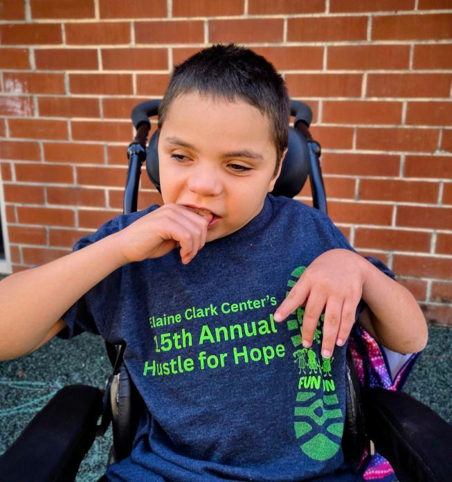 Boy in wheelchair wearing a blue shirt, biting his nails, in front of a brick wall.
