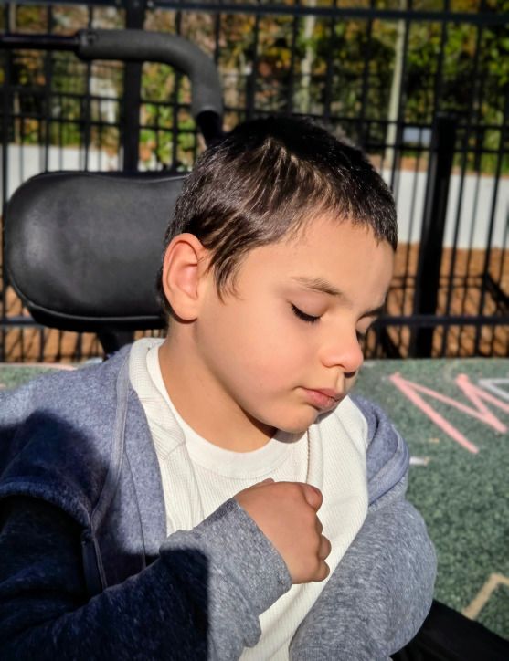 Boy in a wheelchair with eyes closed, wearing a white shirt and gray jacket outdoors in sunlight.