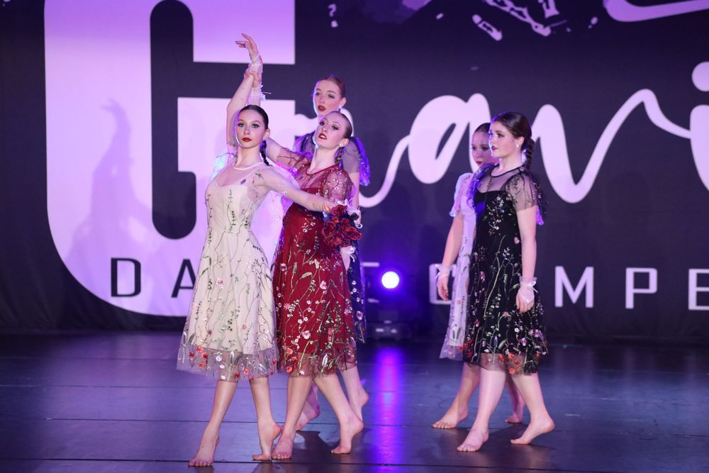 Dancers in formal dresses on stage during a competition. Their expressions and poses suggest graceful performance.
