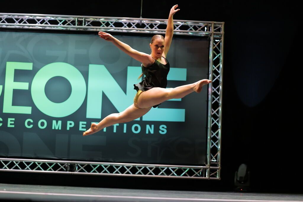 Dancer in mid-air split, arms extended, performing in front of a stage backdrop with a 