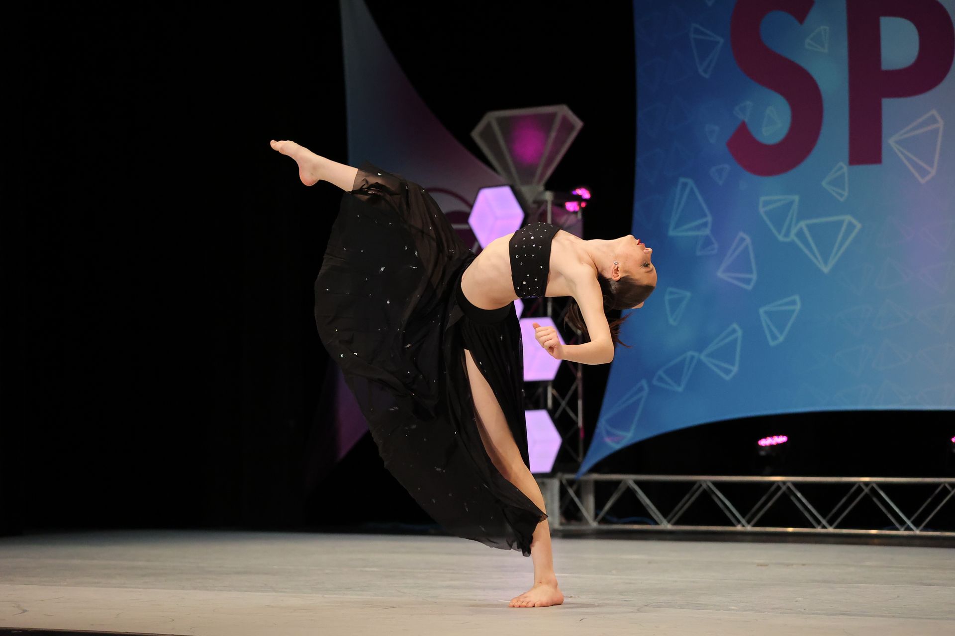 A dancer in a black costume balances in a pose on stage. She is arched backwards with her leg extended.