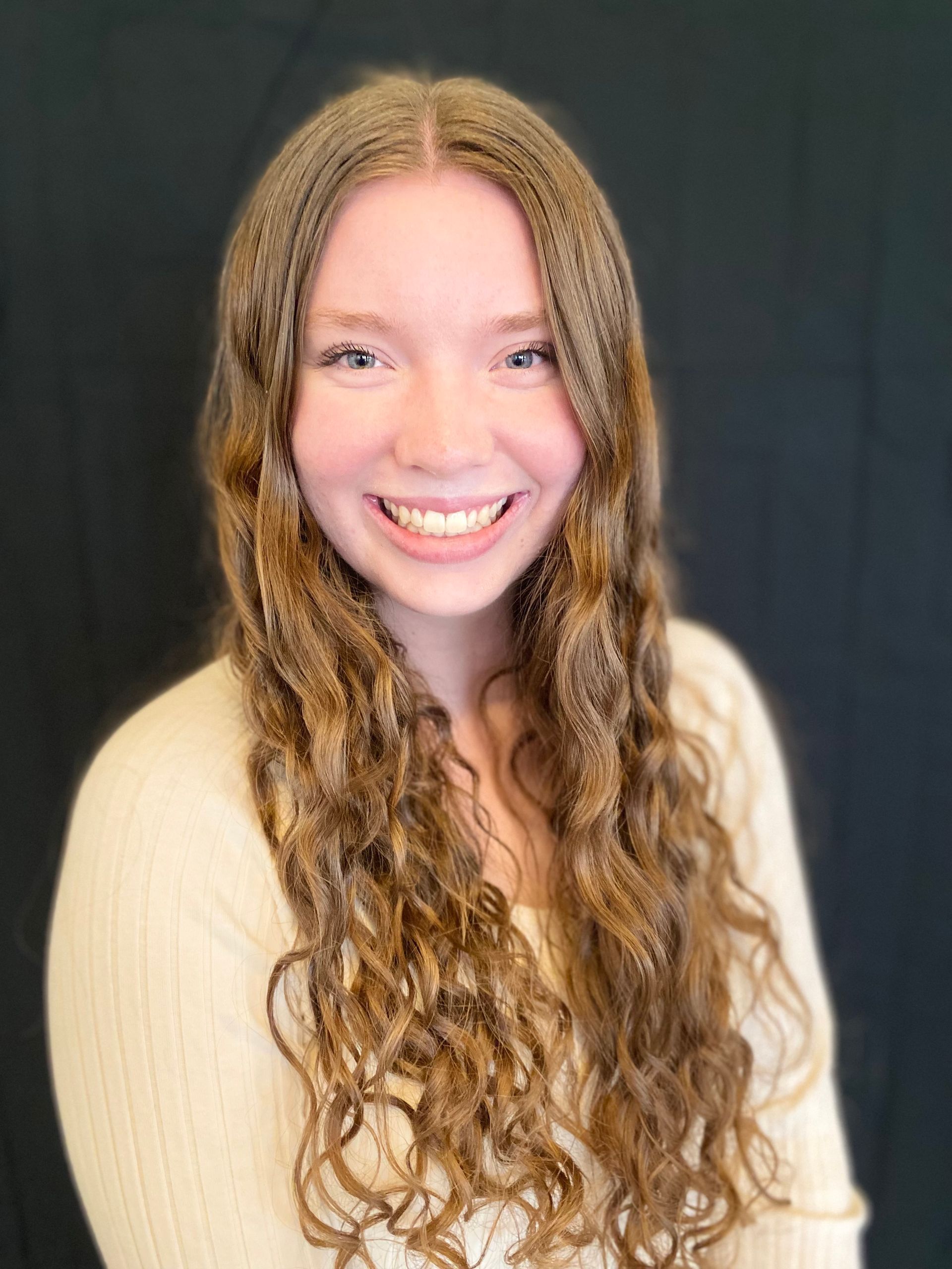 Young woman with long, wavy reddish-brown hair, smiling broadly at the camera. She wears a cream-colored sweater against a dark background.