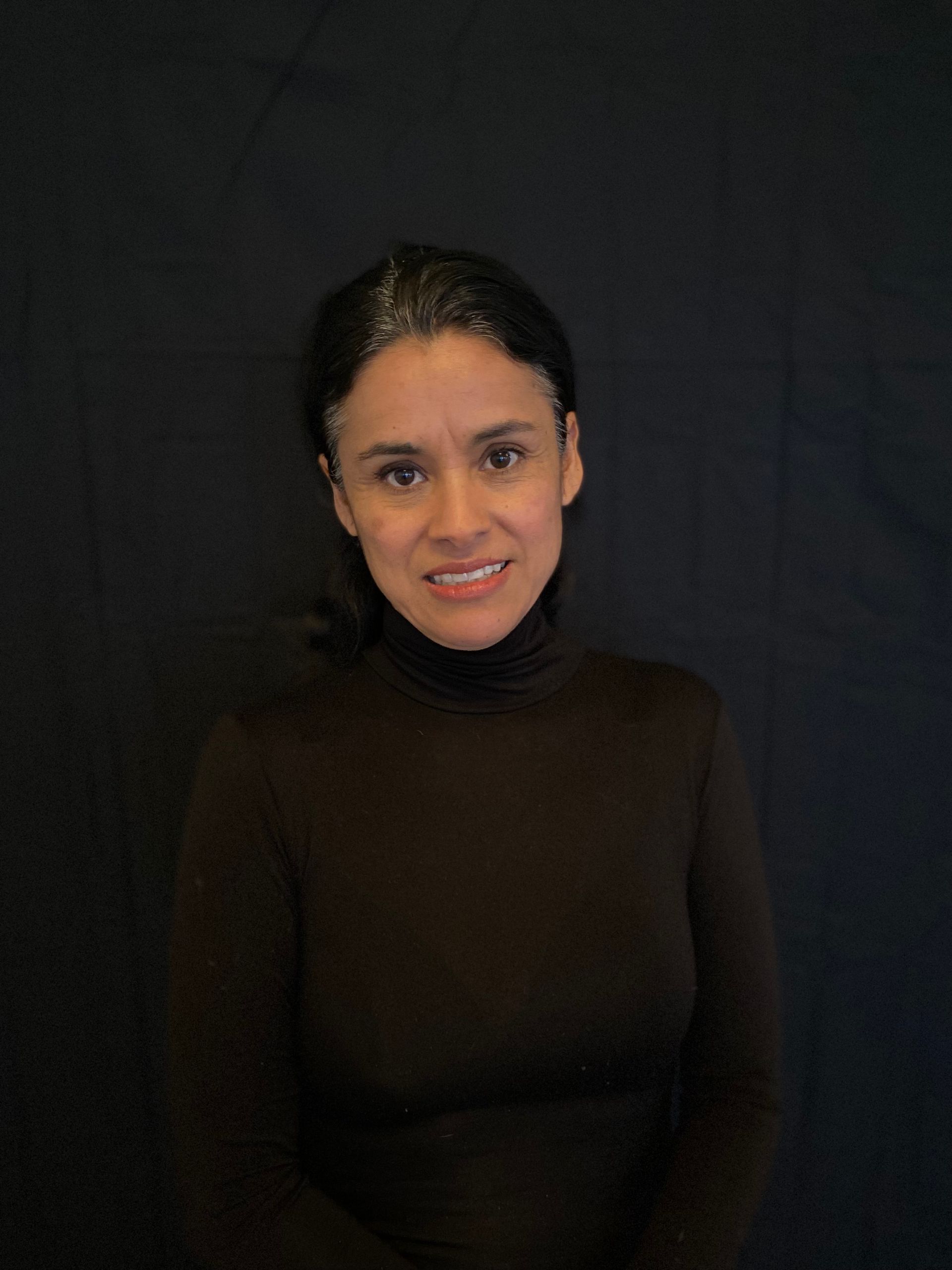 Woman with dark hair in a black turtleneck smiles at the camera against a black backdrop.