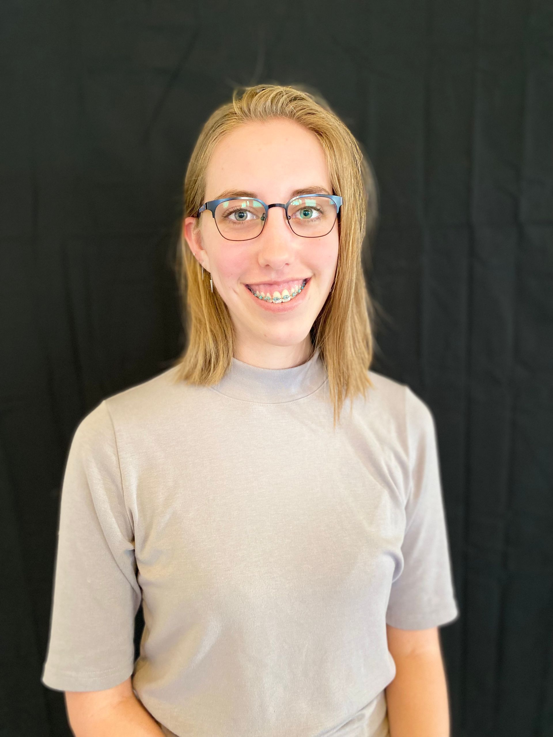 Woman with blonde hair and glasses smiles in front of a black backdrop, wearing a gray, short-sleeved shirt.