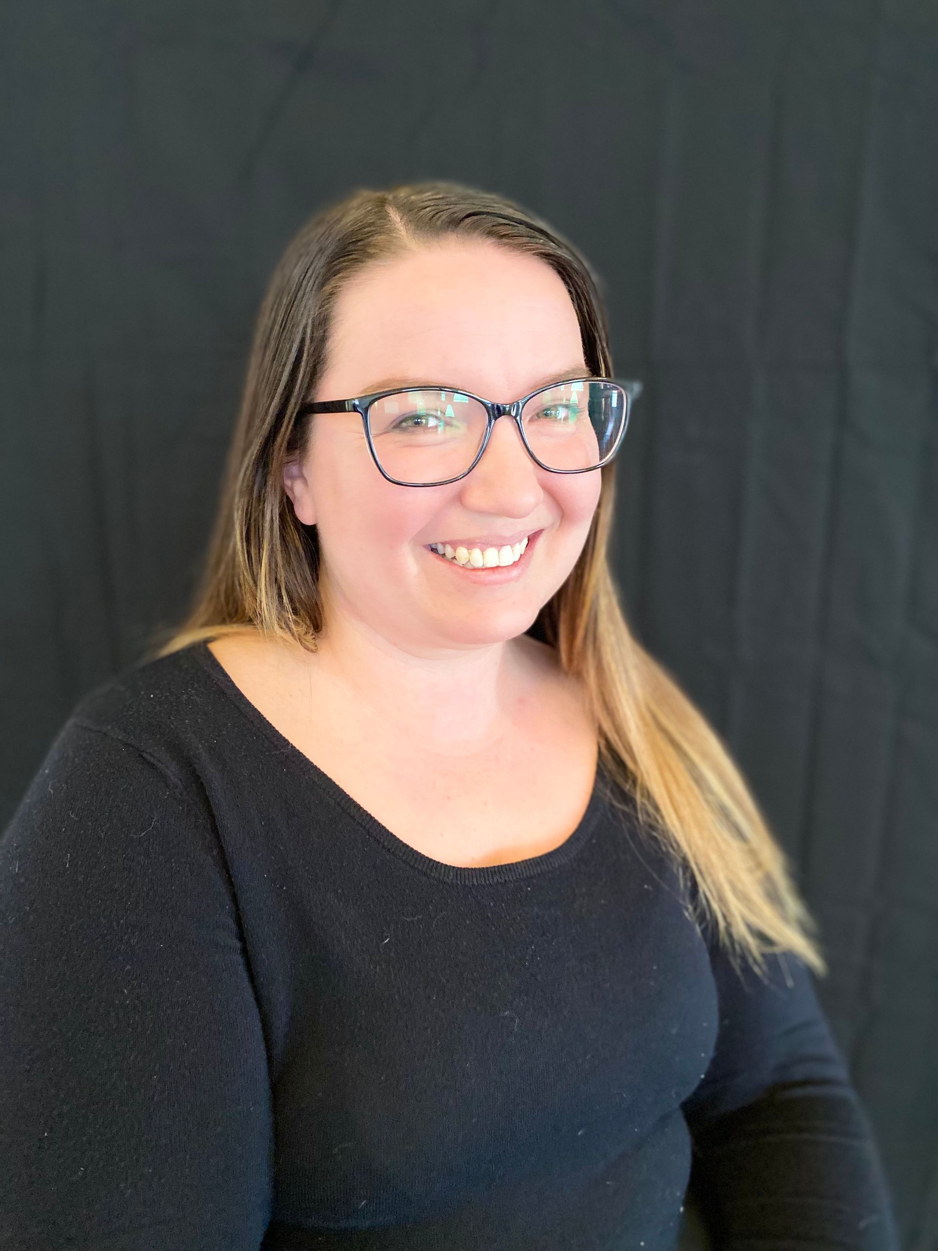 A woman with glasses and long blonde hair smiles at the camera, wearing a black sweater, against a black backdrop.