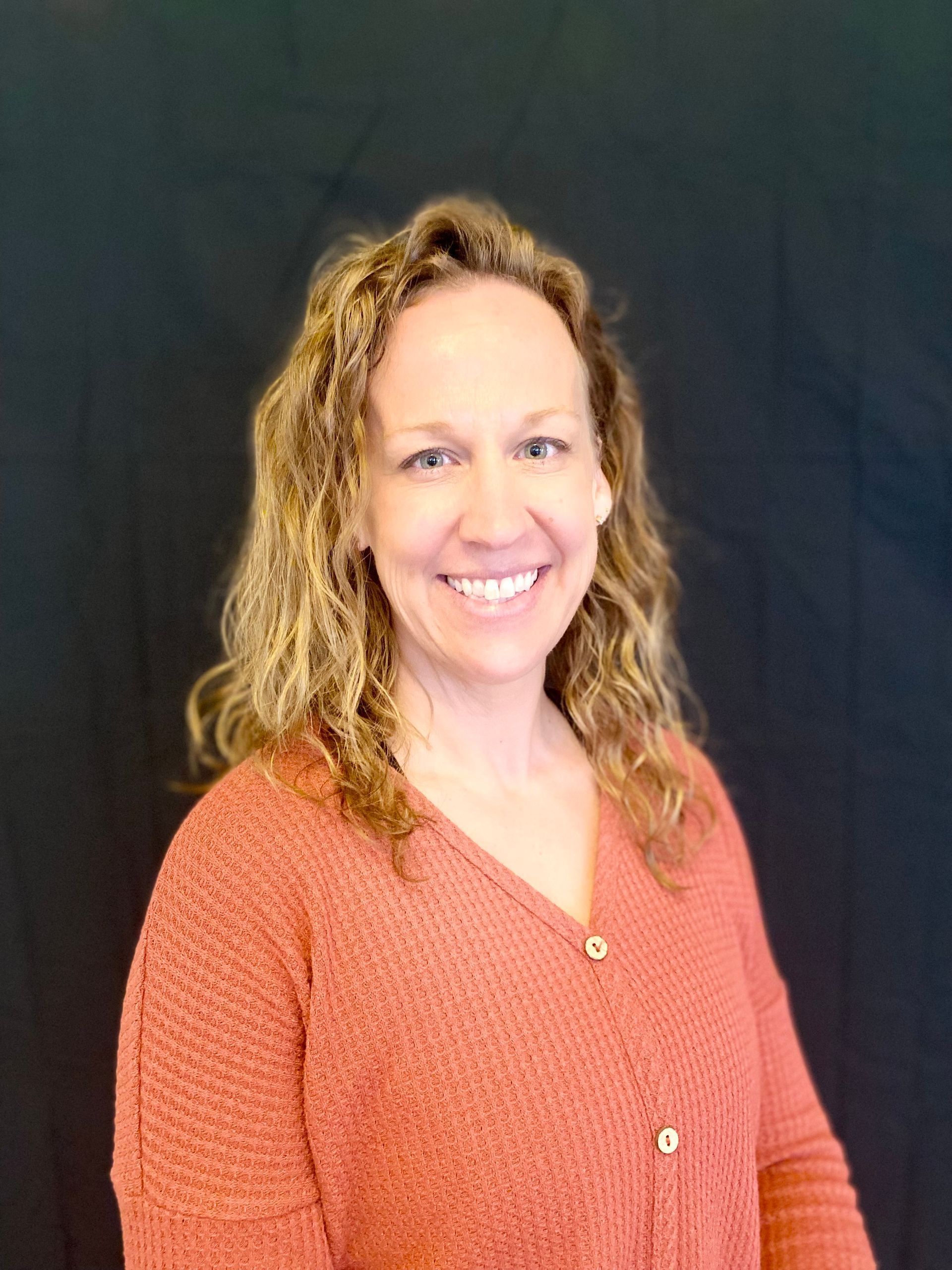 Woman with blonde curly hair smiles, wearing a coral sweater. Black backdrop.